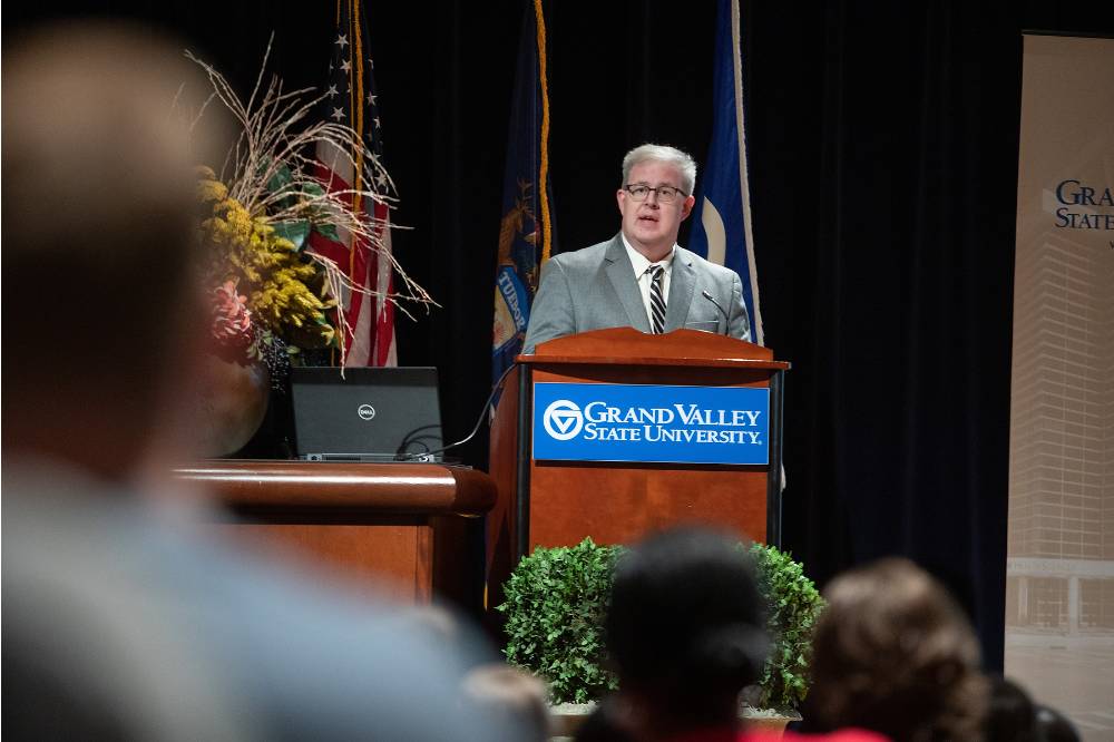 Man stands in focus at a GVSU podium, with people sitting out of focus to the side